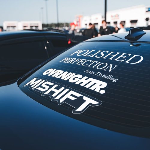 Rear view of black car with custom window decals at an outdoor auto event.