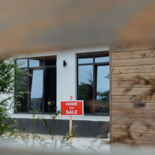 A contemporary home with a 'For Sale' sign in the front yard, framed by greenery.